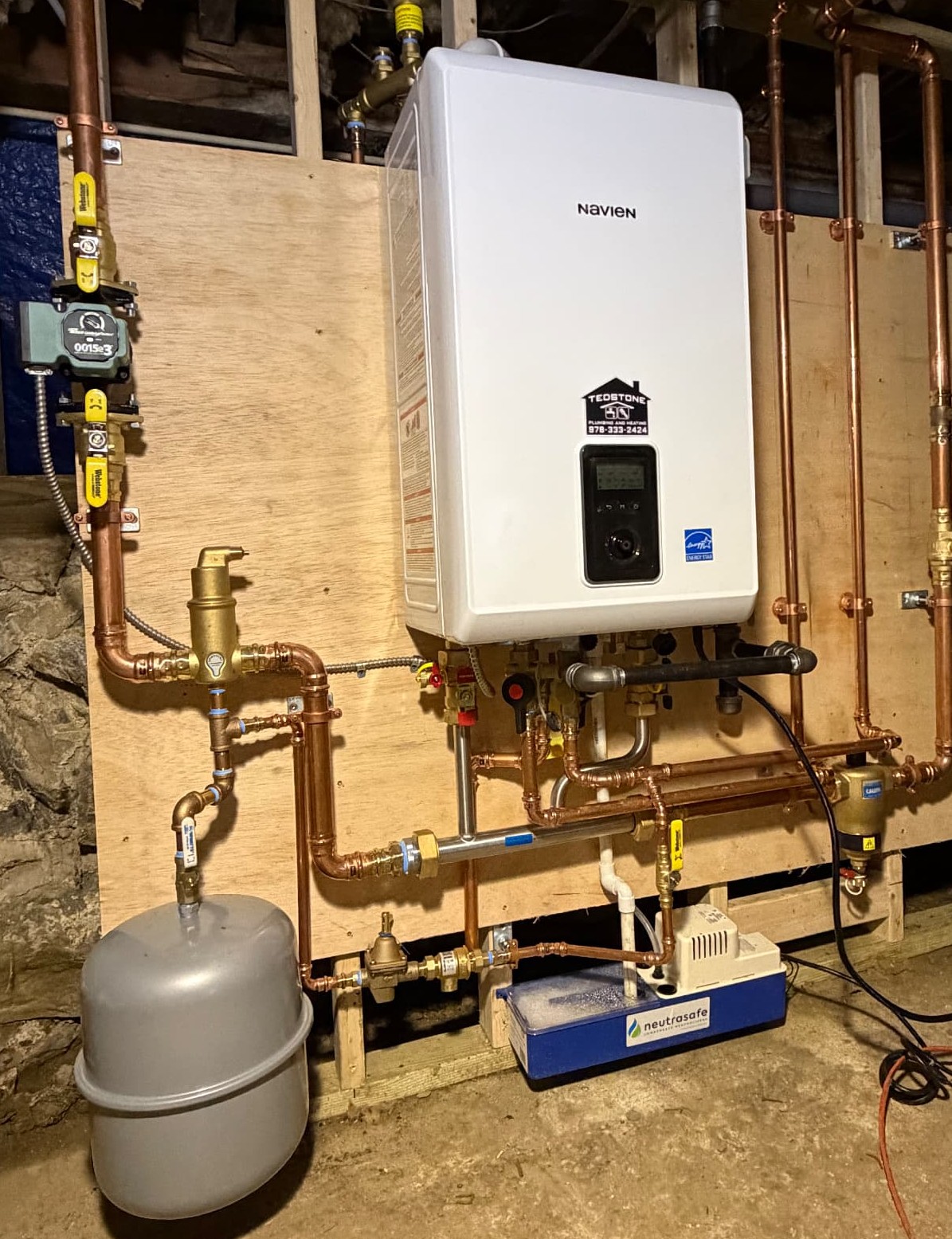 Neat copper pipework with pressed fittings, yellow-handled isolation valves, an expansion tank, a circulator pump and a condensate neutralizer installed on a plywood backer board beneath a wall-mounted Navien unit